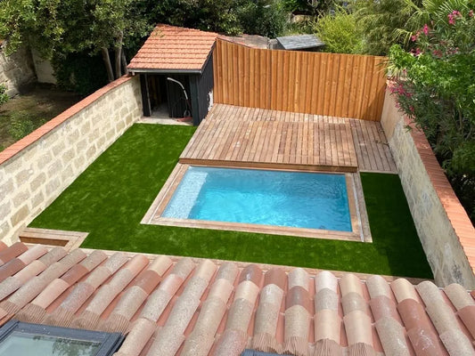 Jardin avec une piscine entourée d’une terrasse en bois et d’une pelouse synthétique, sans la bannière. Vue depuis un toit en tuiles.