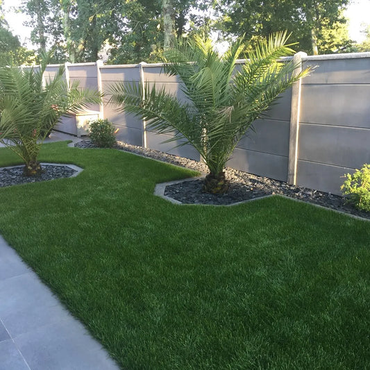 Jardin paysager avec deux palmiers entourés de bordures en pierre, posé sur une pelouse verte parfaitement entretenue, délimitée par une clôture moderne grise