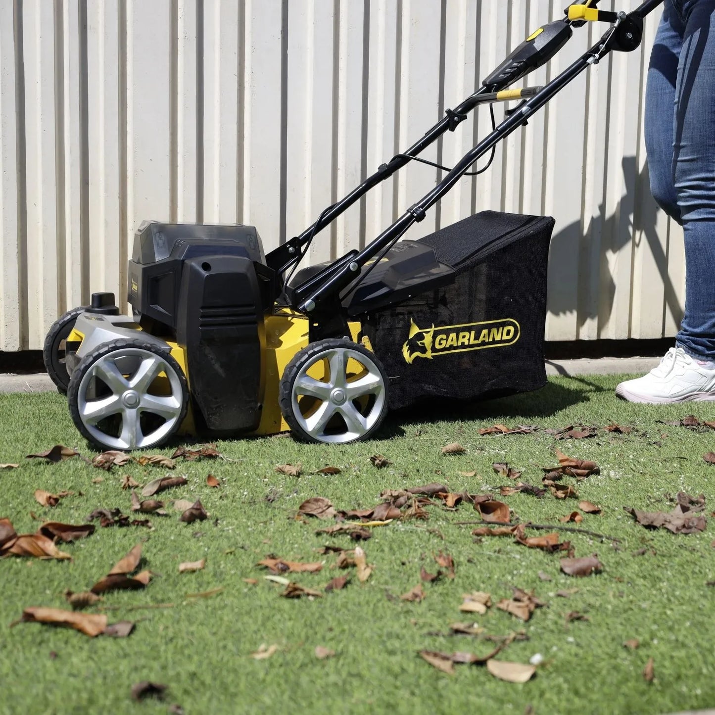 Une brosse électrique Garland Comber 602E en utilisation sur une pelouse synthétique avec des feuilles au sol. La machine est activée pour nettoyer efficacement le gazon.
