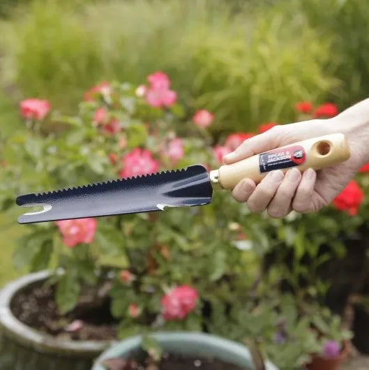 Le couteau désherbeur tenu en main dans un jardin fleuri, illustrant sa facilité d'utilisation pour l'entretien des massifs et des jardinières.