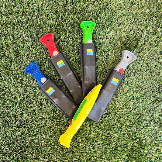 Une collection de cutters multicolores (jaune, rouge, vert, bleu et gris), chacun rangé dans un étui noir. Ils sont disposés en éventail sur une pelouse synthétique.
