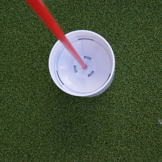 Vue de dessus d'une coupe de golf blanche avec un drapeau rouge inséré au centre, sur une surface de gazon synthétique parfaitement entretenu.