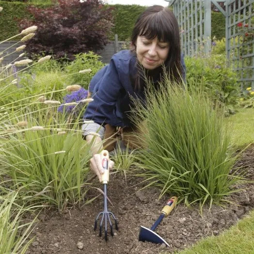 Une jardinière utilise l'outil dans un parterre de jardin, montrant son efficacité pour retirer les mauvaises herbes et ameublir la terre.