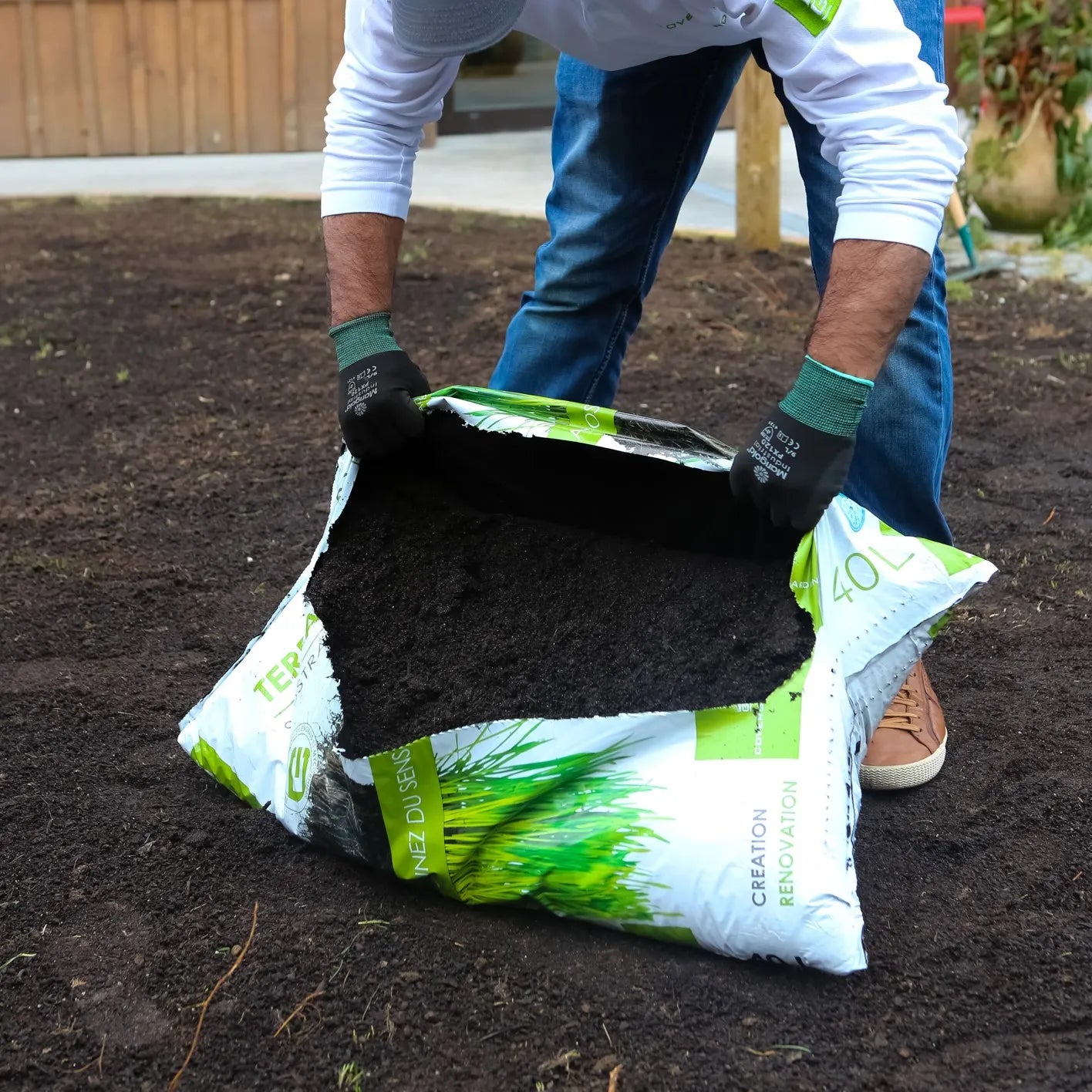 Un homme portant des gants noirs et des chaussures marron ouvre un grand sac de substrat pour gazon sur un sol fraîchement préparé. Le sac est blanc et vert, avec des illustrations d'herbe et le texte "Création Rénovation 40L" visible.