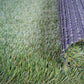 Gazon synthétique vert avec des fibres réalistes, et un morceau enroulé montrant le dessous du tapis noir texturé.