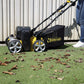 Une brosse électrique Garland Comber 602E en utilisation sur une pelouse synthétique avec des feuilles au sol. La machine est activée pour nettoyer efficacement le gazon.