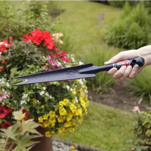  Le couteau désherbeur tenu par une main dans un jardin fleuri, illustrant son utilisation pratique pour l'entretien des espaces verts.