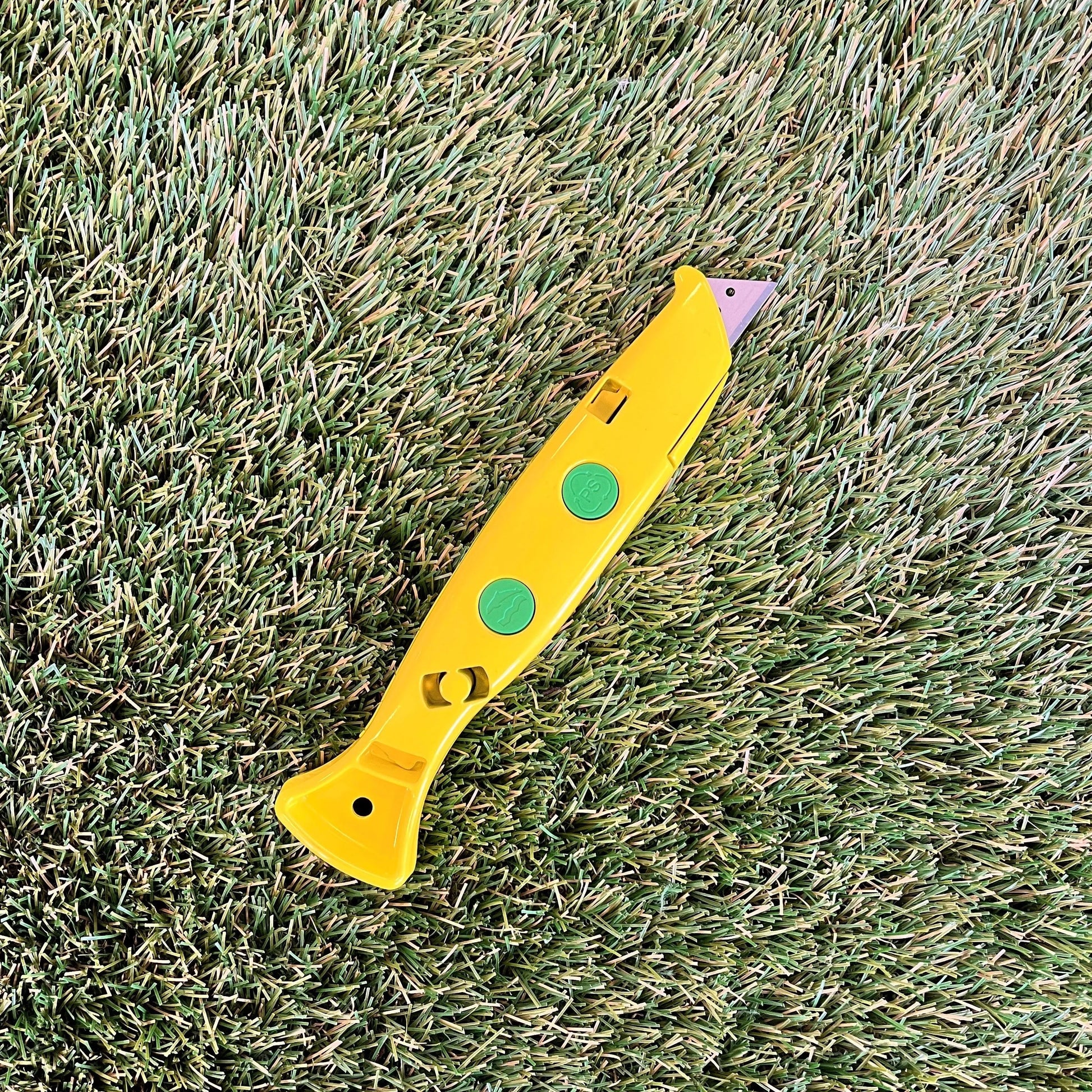 Un cutter jaune vif avec des boutons verts posé sur une pelouse synthétique. Son design ergonomique et sa lame rétractable sont conçus pour des coupes précises.