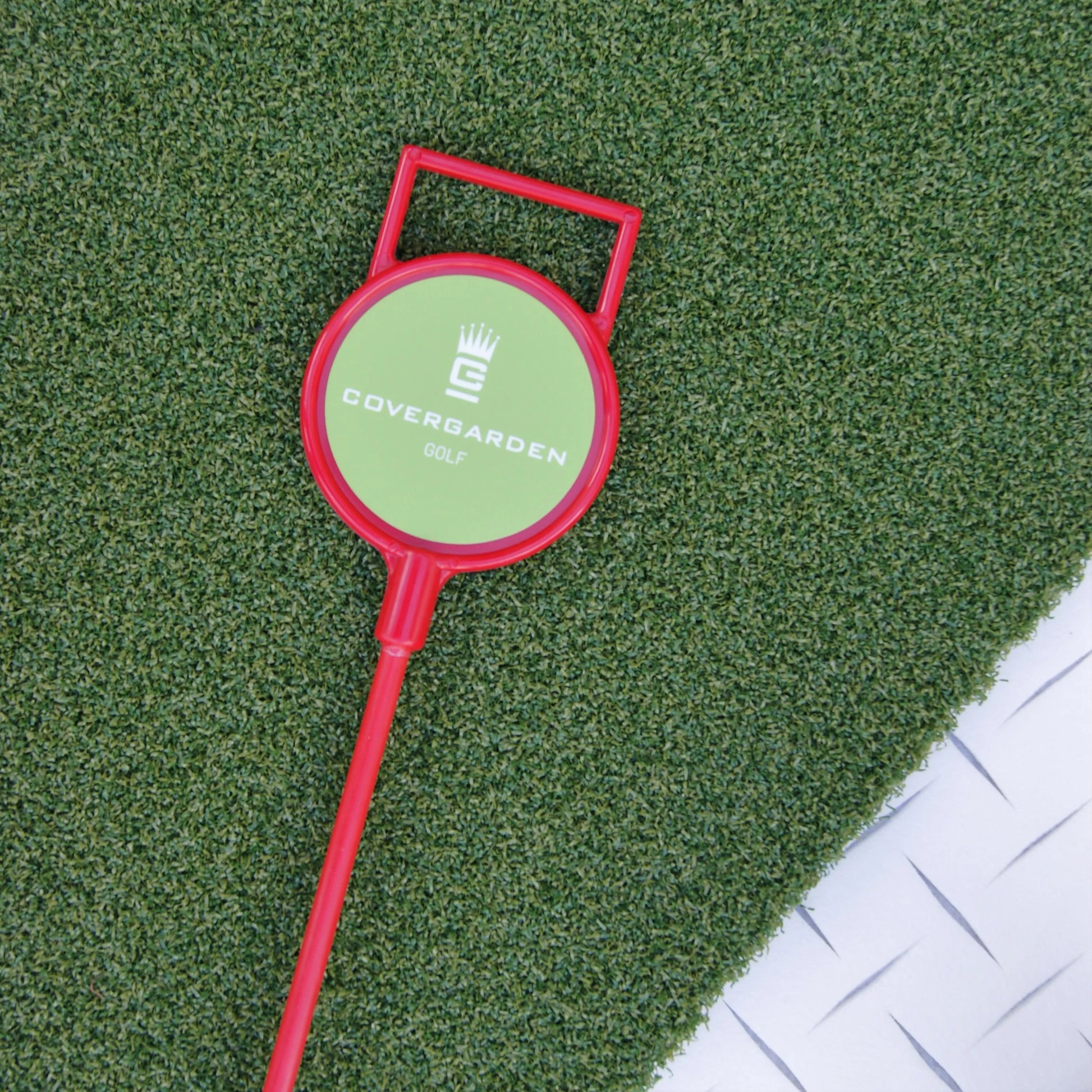 Marqueur de golf rouge avec poignée, portant le logo 'COVERGARDEN GOLF', posé sur une surface de gazon synthétique vert.