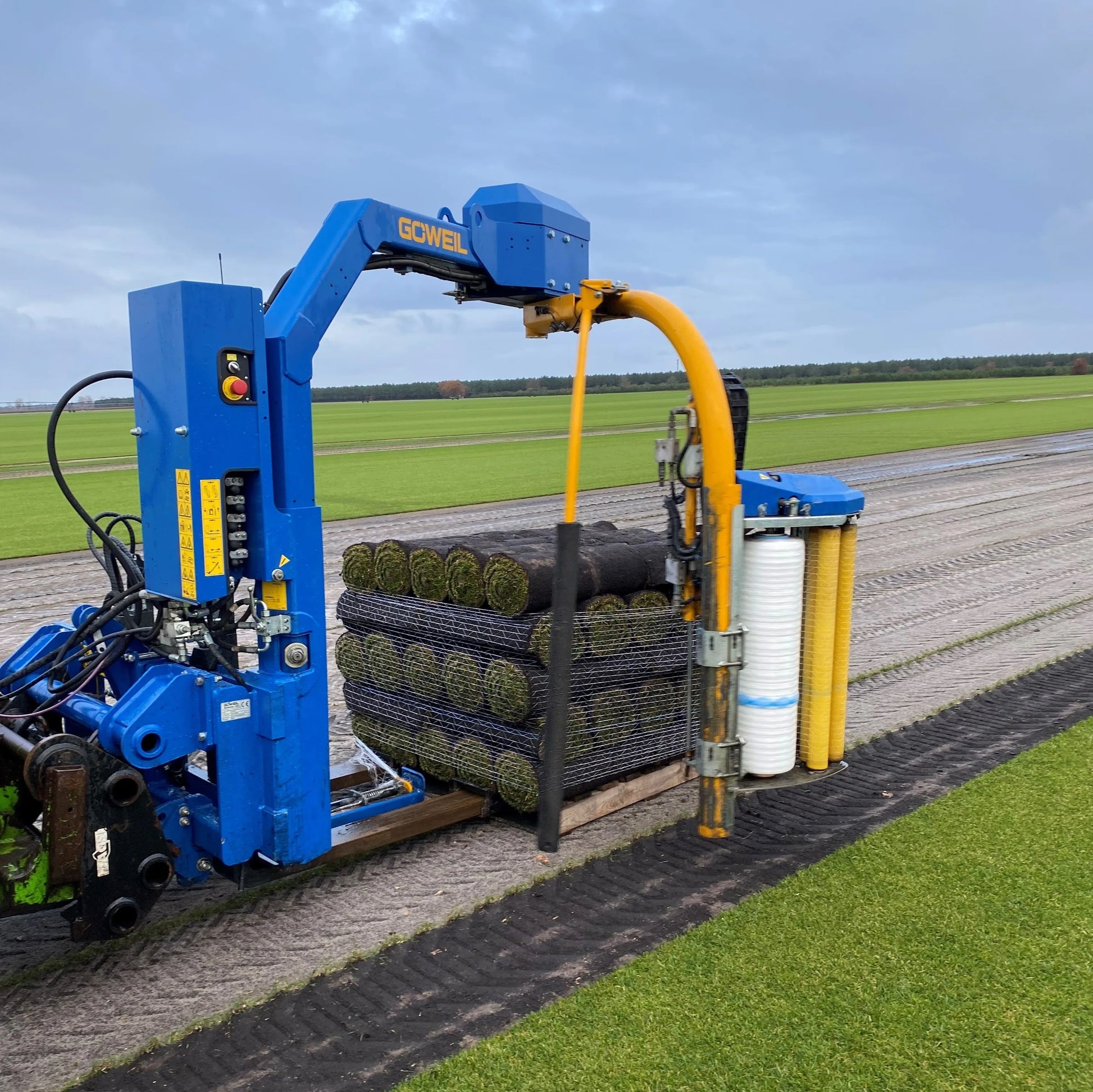 Machine agricole bleue de type récolteuse avec des rouleaux de gazon fraîchement enroulés. La machine est posée sur un champ, avec des rangées de gazon bien entretenues à l'arrière-plan.